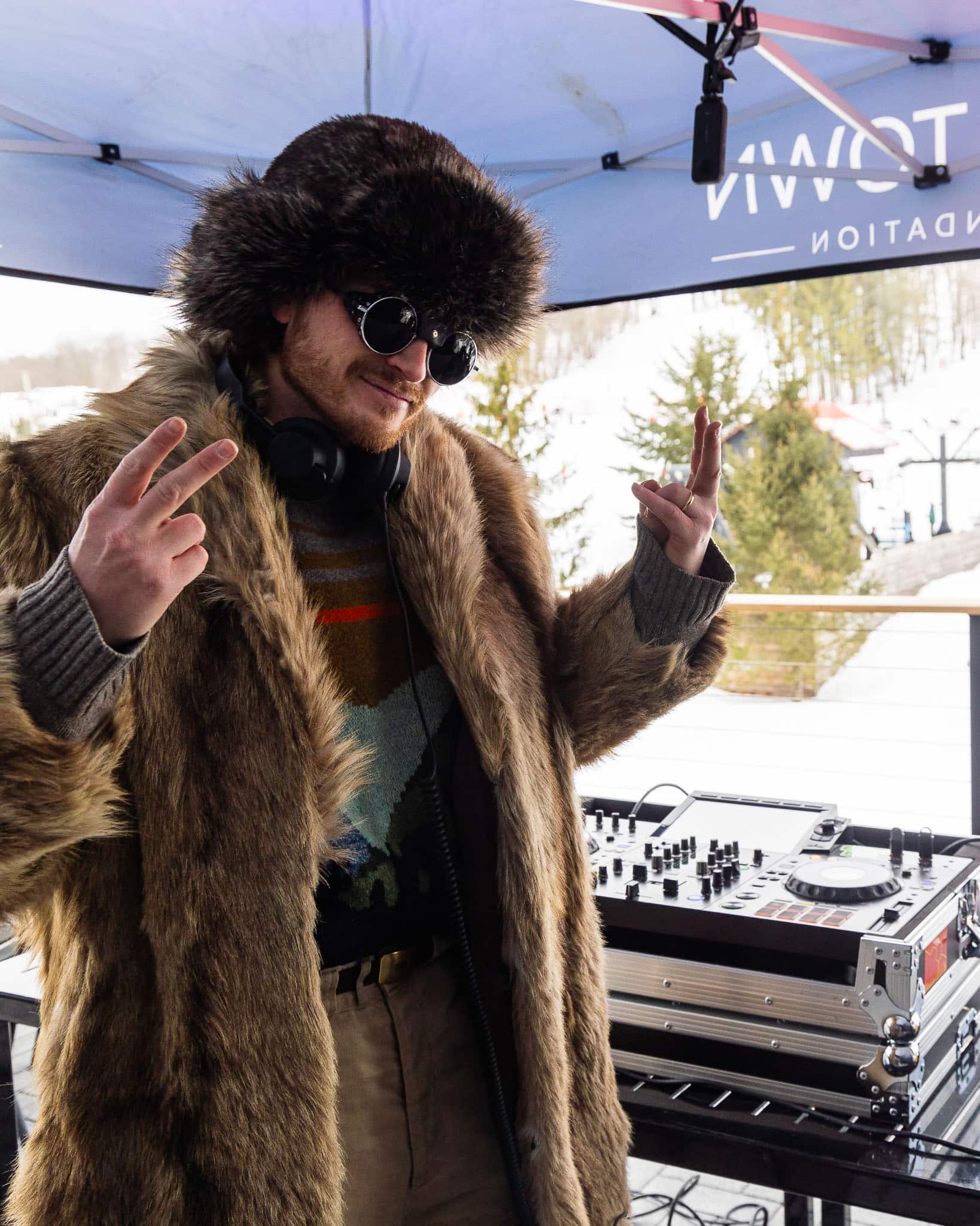 DJ on the deck wearing a fur coat and hat, throwing up peace signs