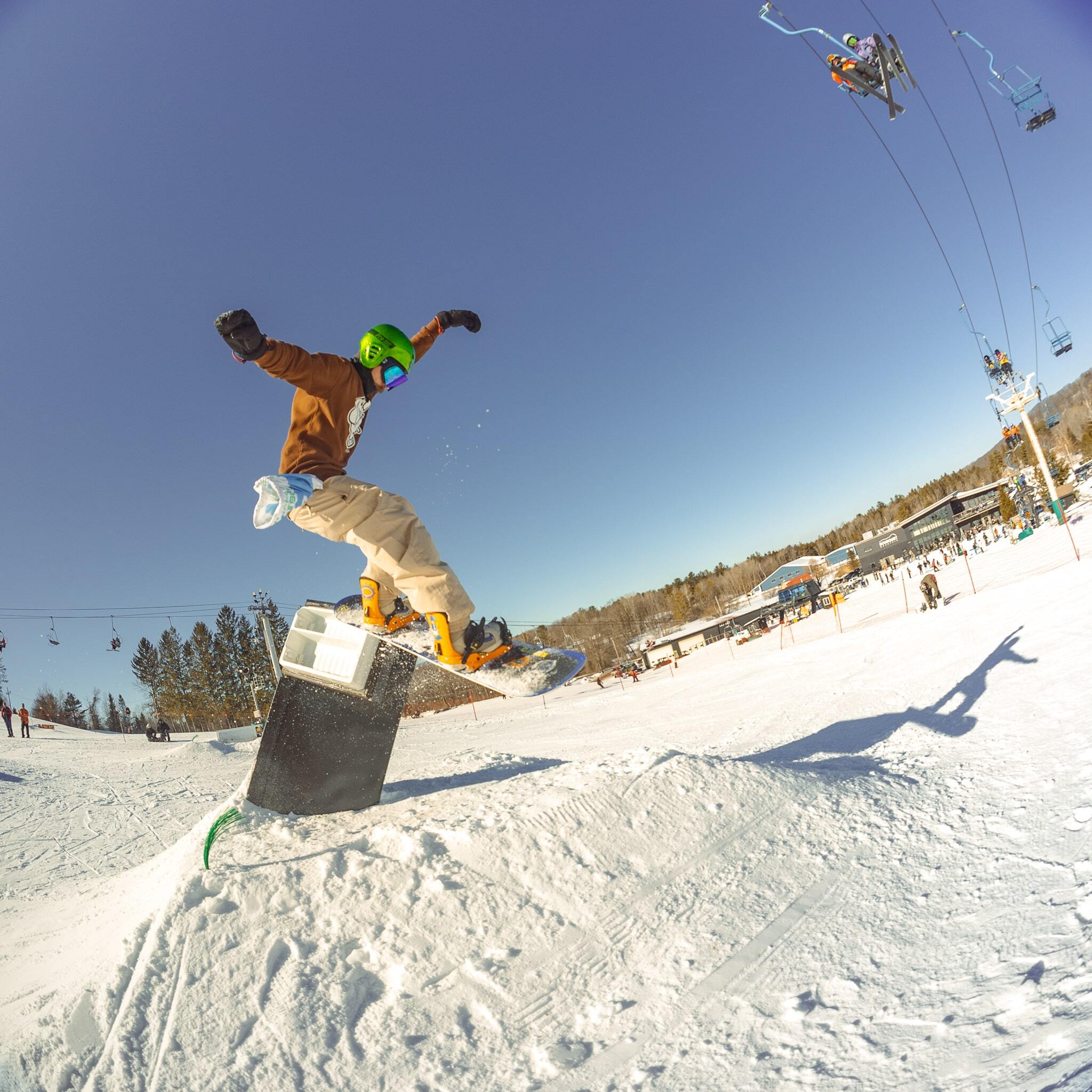 A snowboarder in a brown jacket, beige pants, and green helmet launches off a box feature in the terrain park on a clear blue-sky day, with the ski lift, lodge, and base area of a mountain resort visible in the background.