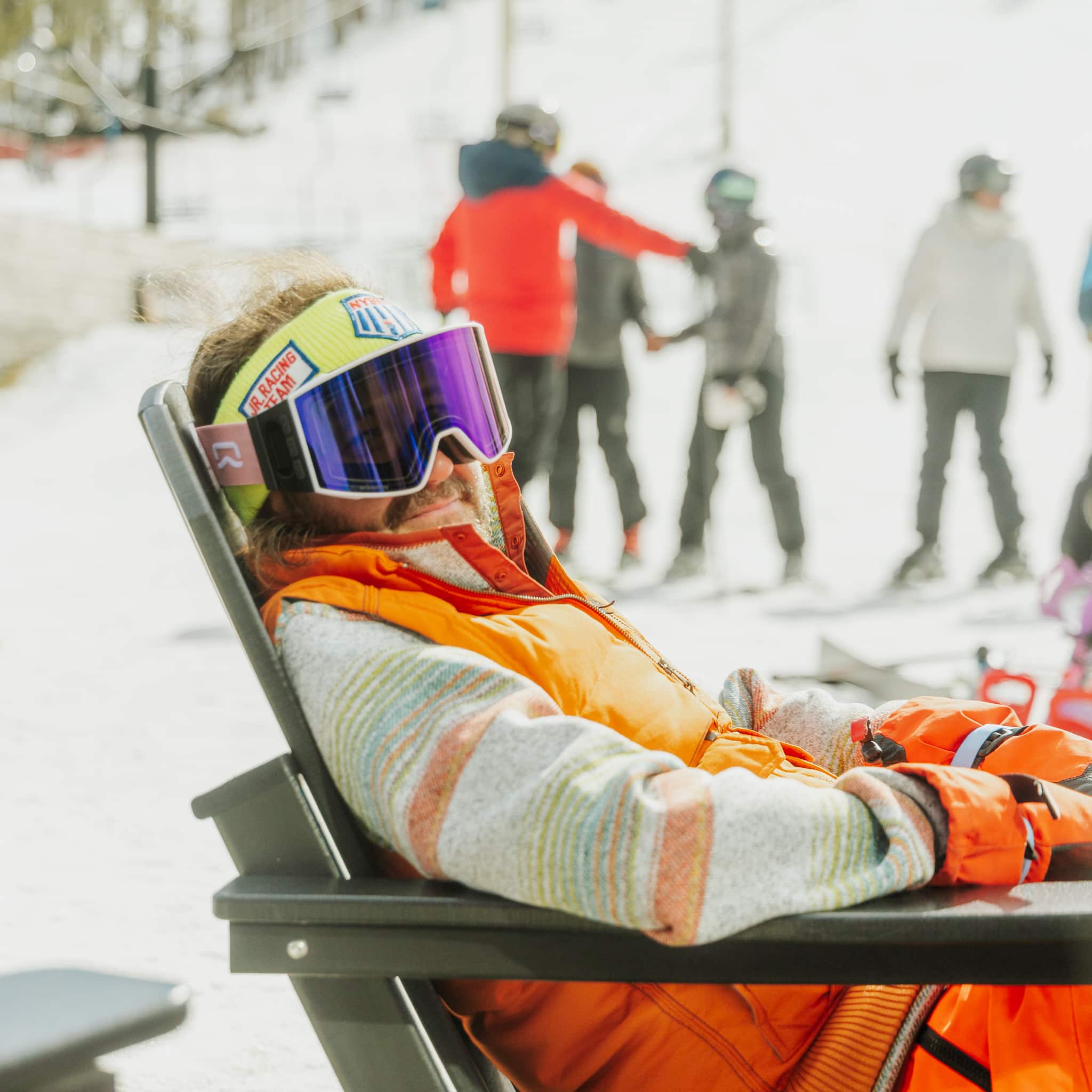 A bearded man in an orange ski jacket and colorful striped sweater reclines in an Adirondack chair on a sunny day, wearing oversized purple ski goggles and a yellow Jr. Racing Team headband, with skiers visible on the slope behind him.