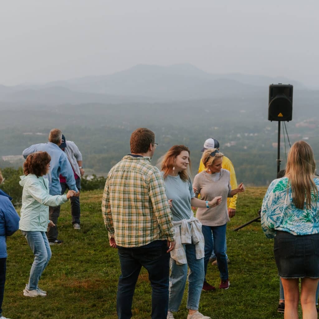 Group of people standing in a grassy field with hazy hills in the background and a speaker on a stand at the right.