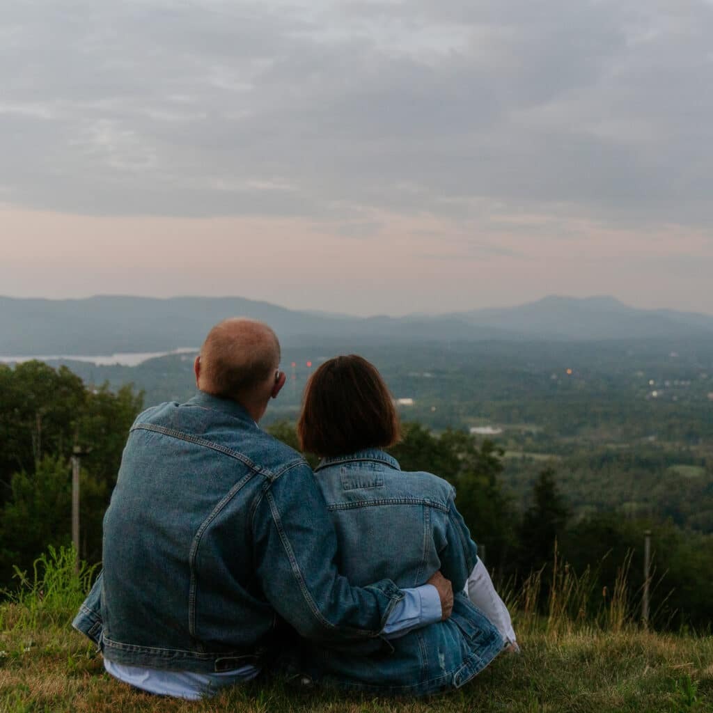 Two people sit on a grassy hillside with their backs to the camera, wearing denim jackets, overlooking a valley and distant mountains under a cloudy sky.