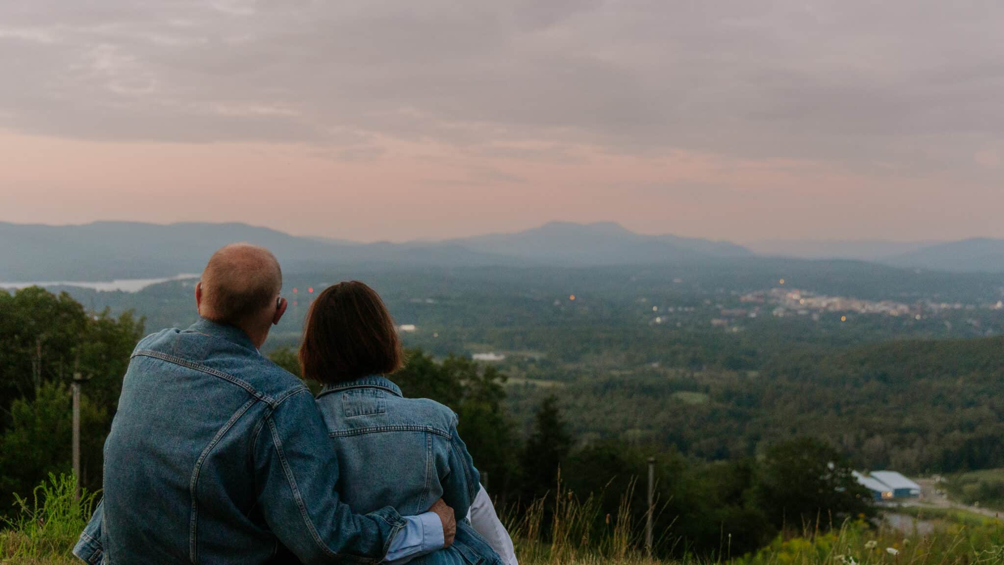 Couple in denim jackets sitting on a grassy hillside, cuddled together and looking out over a vast valley with distant mountains at sunset.