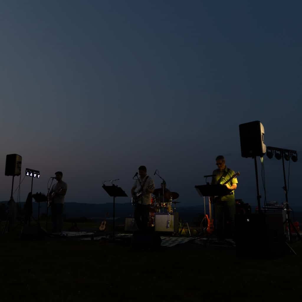 Outdoor concert at dusk with silhouettes of musicians on a stage, guitars and drums visible under stage lights.