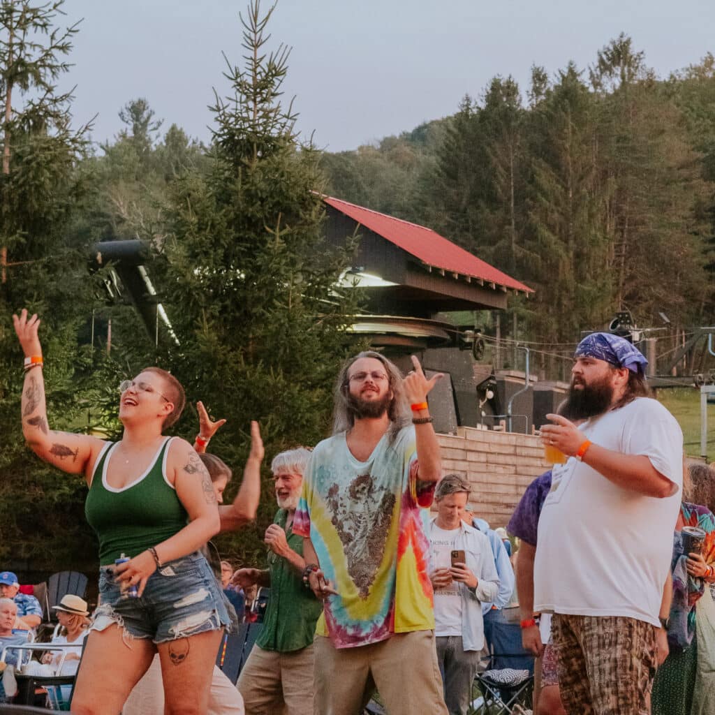 Group of people cheering at an outdoor festival, some with arms raised and colorful clothing, trees in the background