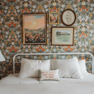 White metal-bed bedroom with bold floral wallpaper, framed art above, and a small pillow that reads 'Bit of a mess but doing my best'.