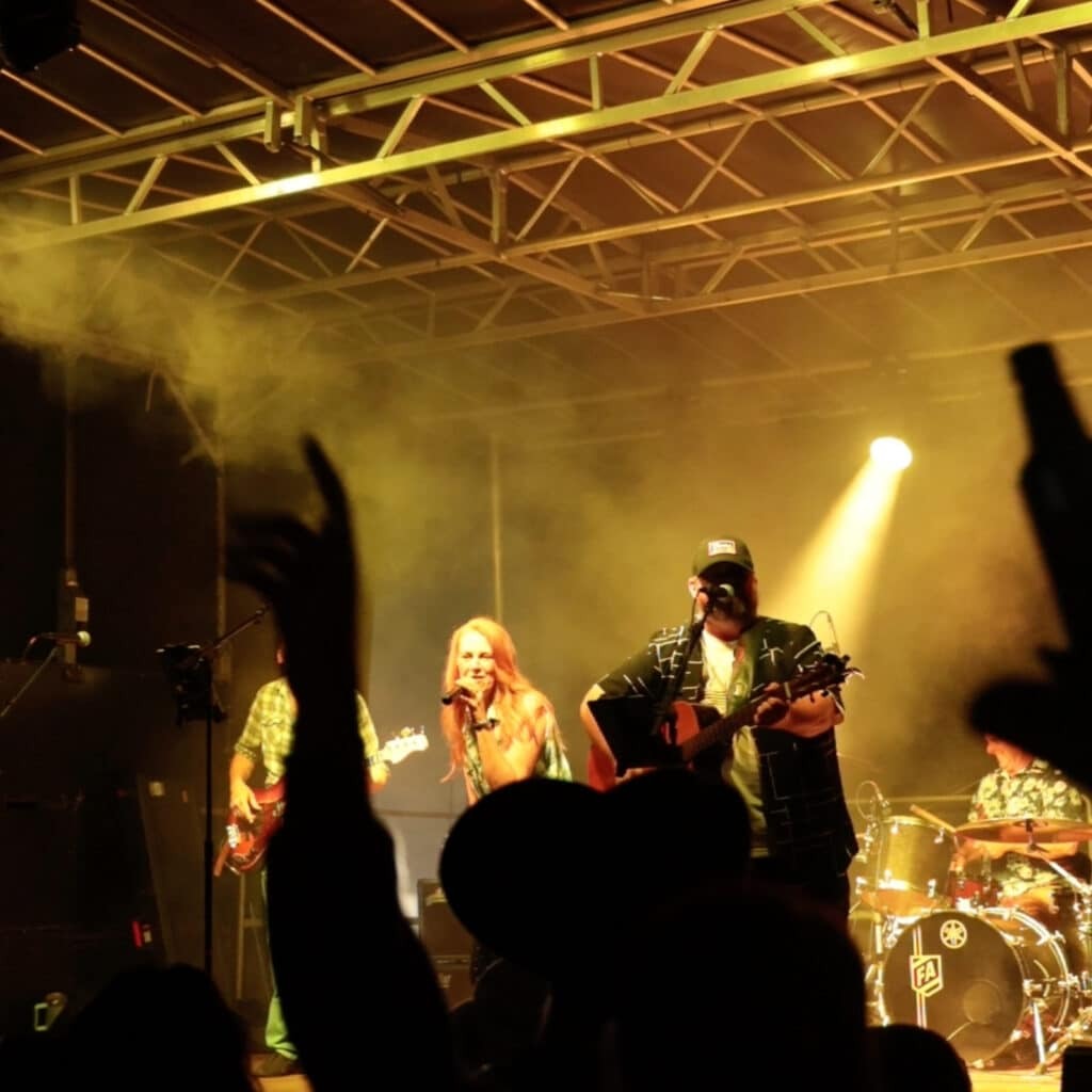 Live band performs on stage with guitarists and a vocalist, yellow stage lights and smoke, crowd silhouettes with raised hands in the foreground