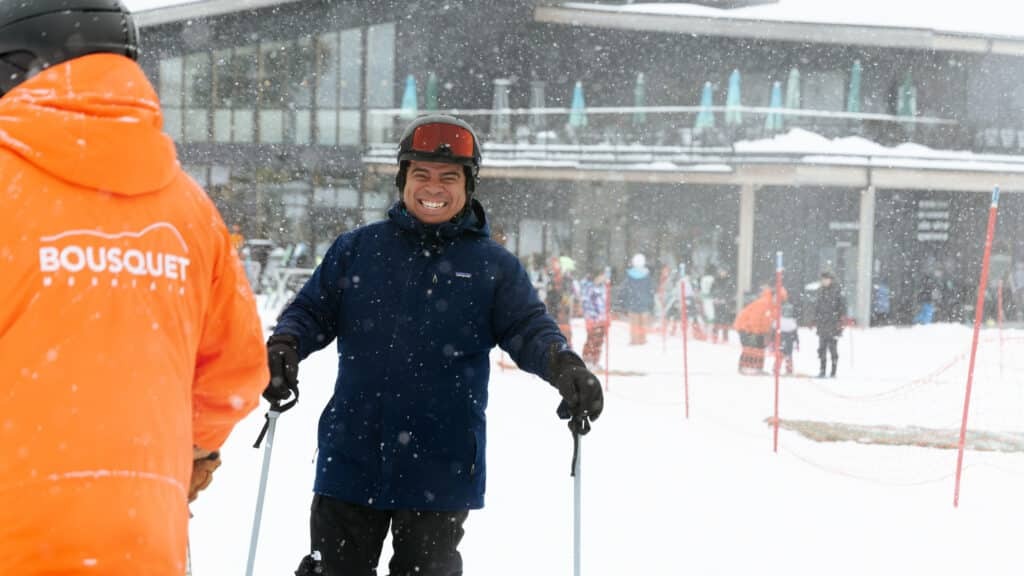 Smiling man in a navy ski jacket and helmet holds ski poles while skiing in heavy snowfall, with an orange-jacketed staff member nearby on a snowy slope.