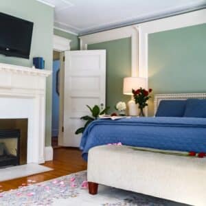 Hotel bedroom with green walls, a blue quilted bed, white fireplace, and an open door to a hallway; roses and petals scattered on the floor.